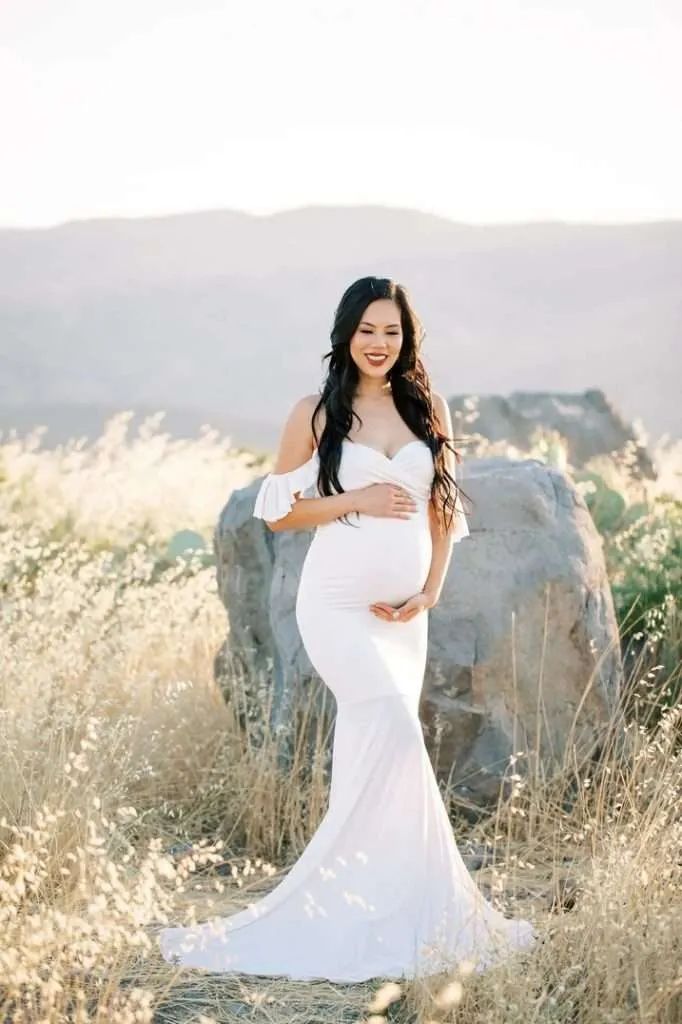 A pregnant woman in a white dress stands outdoors in a field with tall dry grass and large rocks, holding her belly and smiling, captured beautifully by a maternity photographer in Phoenix.