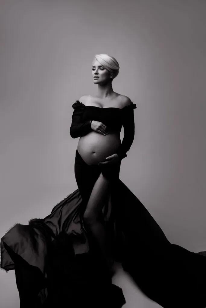 A pregnant woman in a long, flowing off-shoulder dress stands in a Phoenix studio, cradling her belly with one hand and looking to the side—an elegant moment captured through studio maternity photography.
