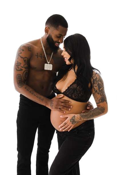 A shirtless, tattooed man gently holds the belly of a pregnant woman in a black lace top and pants; both stand close together against a white backdrop, capturing an intimate moment in studio maternity photography Phoenix style.