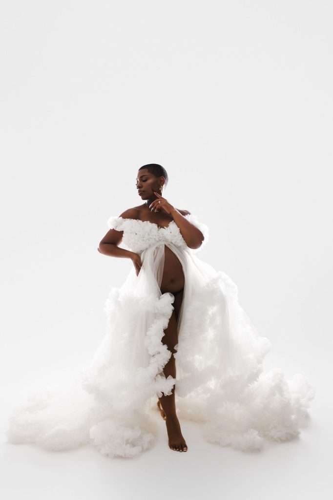 A pregnant woman poses in a sheer, ruffled white gown against a plain white background, showcasing the elegance of studio maternity photography in Phoenix with one hand on her hip and the other at her neck.