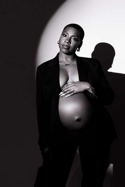 Black and white photo by a maternity photographer Phoenix of a pregnant woman in a dark blazer, posing against a wall with one hand on her exposed belly and the other on her chest.