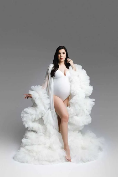 A pregnant woman in a white bodysuit and voluminous tulle robe poses against a plain gray background, capturing the elegance often highlighted by a skilled maternity photographer in Phoenix.