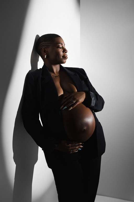 A pregnant woman in a black blazer stands against a white wall in partial shadow, with one hand on her belly and the other resting on her chest—a striking example of studio maternity photography in Phoenix.