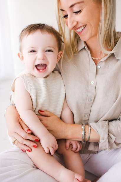 Woman holding a smiling baby on her lap, both in light-colored clothing, in a bright indoor setting—contact maternity photographer Phoenix for precious moments like these.