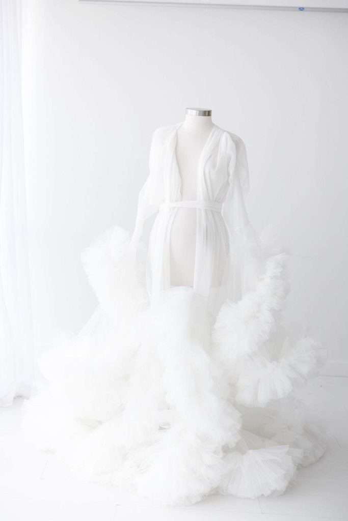 A white mannequin displays a sheer, floor-length white robe with voluminous ruffled tulle accents, set against a bright white background.