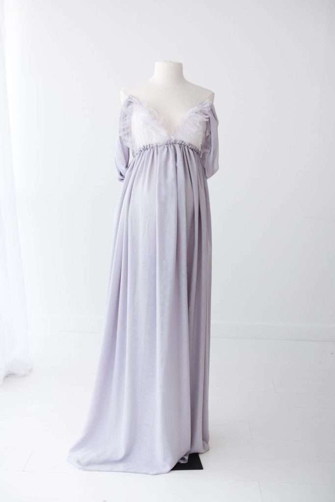 A white mannequin displays a floor-length, off-the-shoulder lavender maternity gown with lace trim on the bodice, set in a minimal white room.