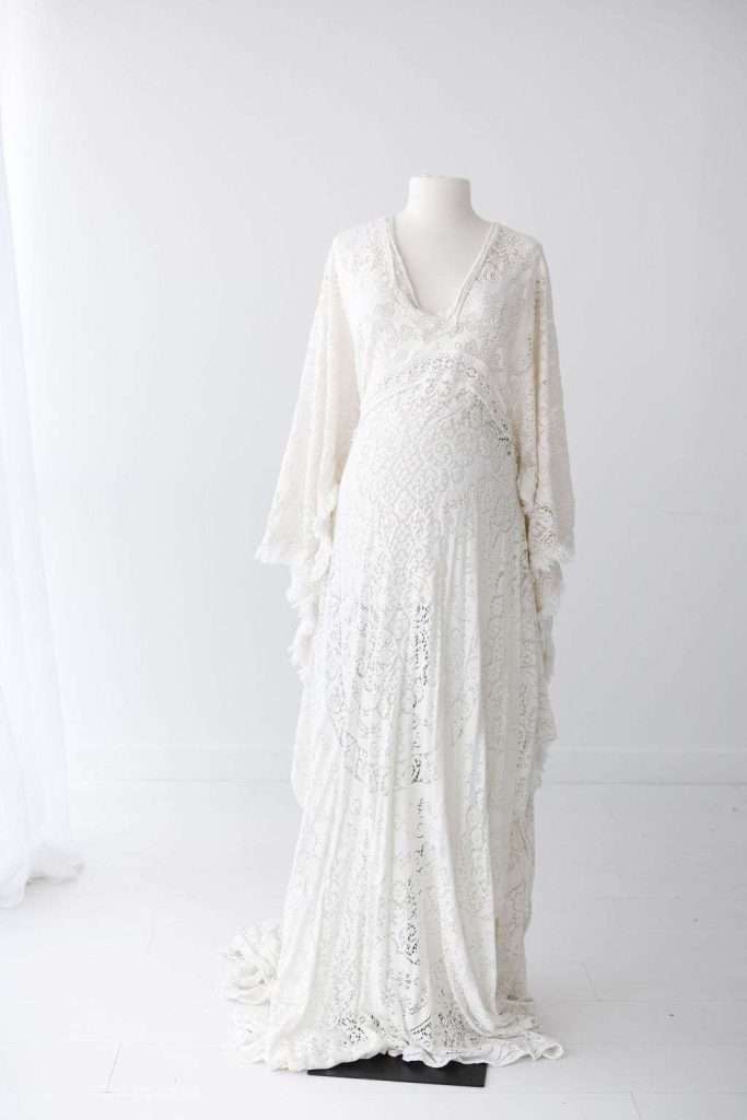 A white lace maternity gown with long sleeves is displayed on a dress form in a minimalist, white room.