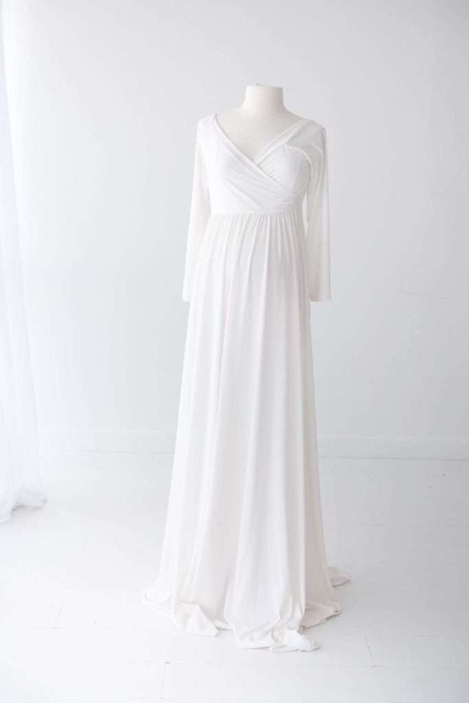 A white, floor-length, long-sleeved dress with a V-neckline displayed on a headless dress form in a minimalist, light-filled room.