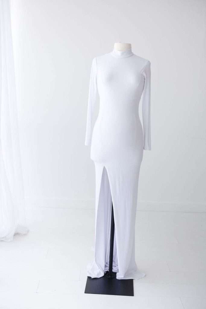 A white, long-sleeve, floor-length gown with a high neckline and front slit displayed on a mannequin in a minimalist white setting.
