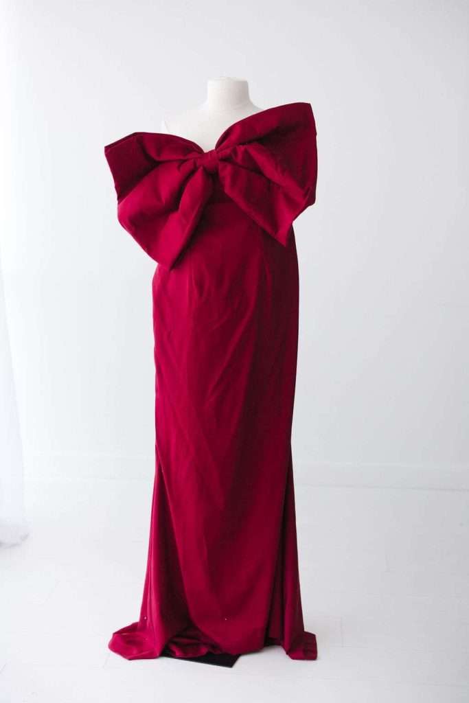 A mannequin displays a floor-length, strapless red gown with a large bow detail at the chest, set against a plain white background.