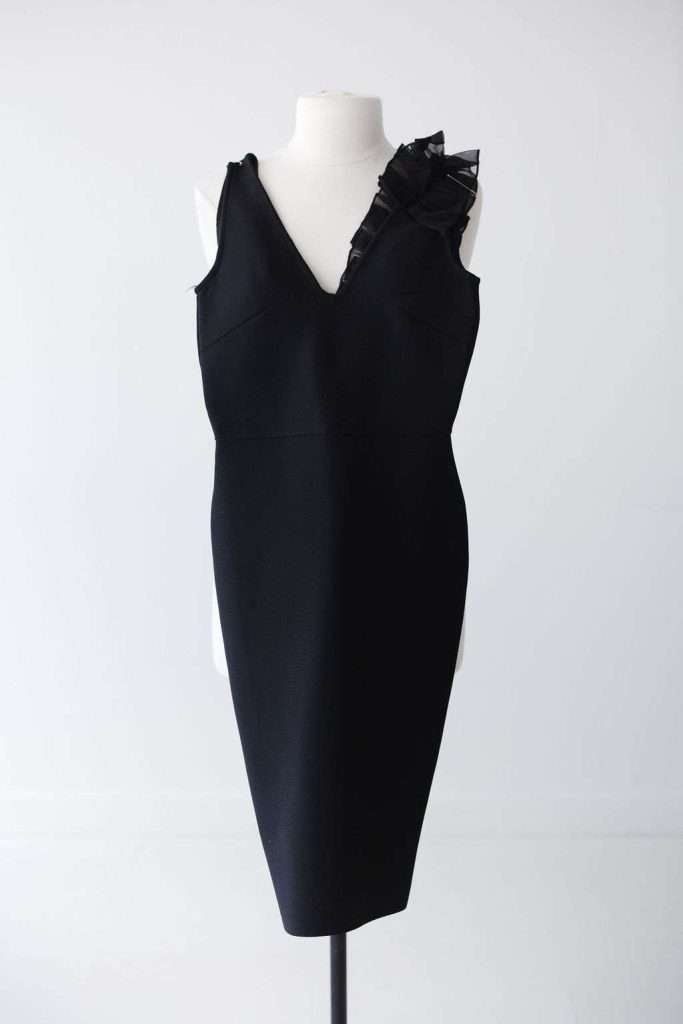 Sleeveless black knee-length dress with a V-neckline and a decorative black ruffle on one shoulder, displayed on a mannequin against a plain background.
