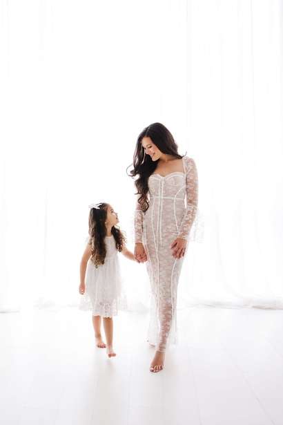 A woman in a long, sheer white lace dress holds hands with a young girl in a short white dress, standing together in a bright, white room—contact a maternity photographer Phoenix to capture moments like these.