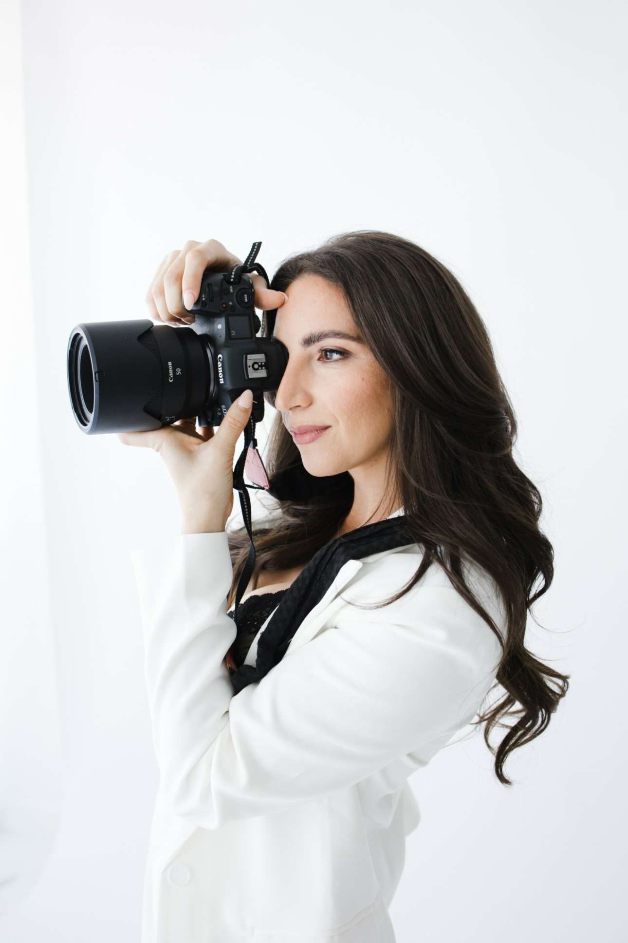 A woman with long dark hair, dressed in a white blazer, holds a camera up to her face as she prepares to take a photograph against a plain white background.