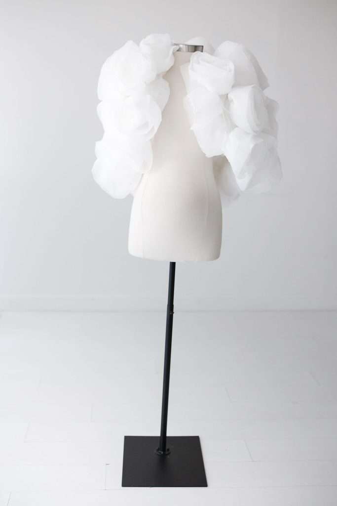 A white dress form displays a garment with large, fluffy, white organza ruffles on the shoulders. The background is plain and minimalistic.