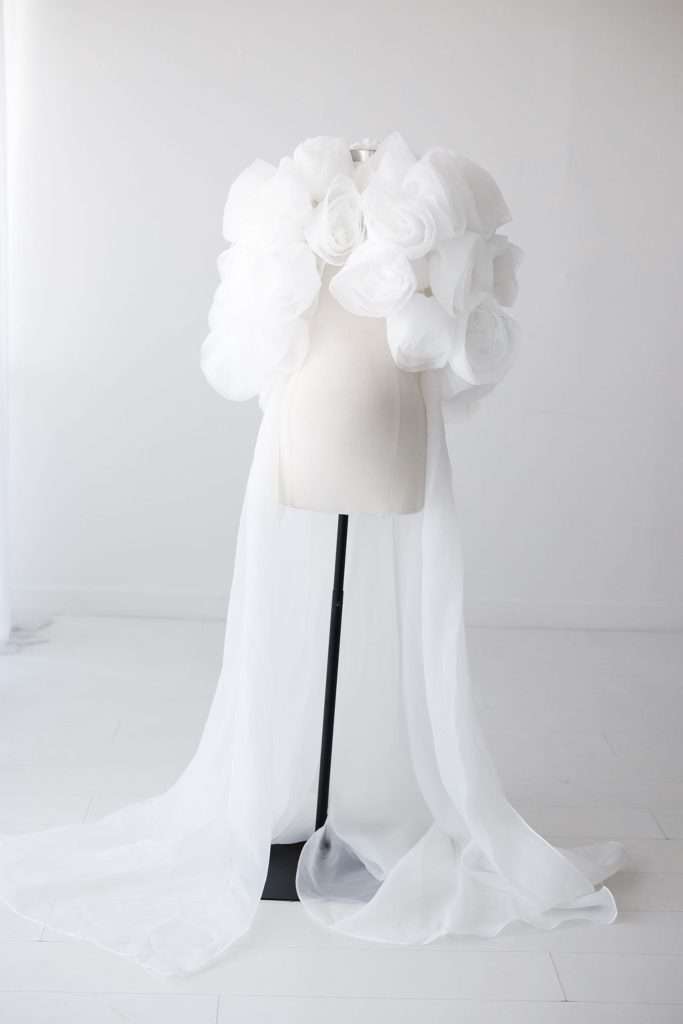 A white dress form displays a garment with large sheer fabric flowers on the shoulders and long flowing sheer white fabric draped to the floor.