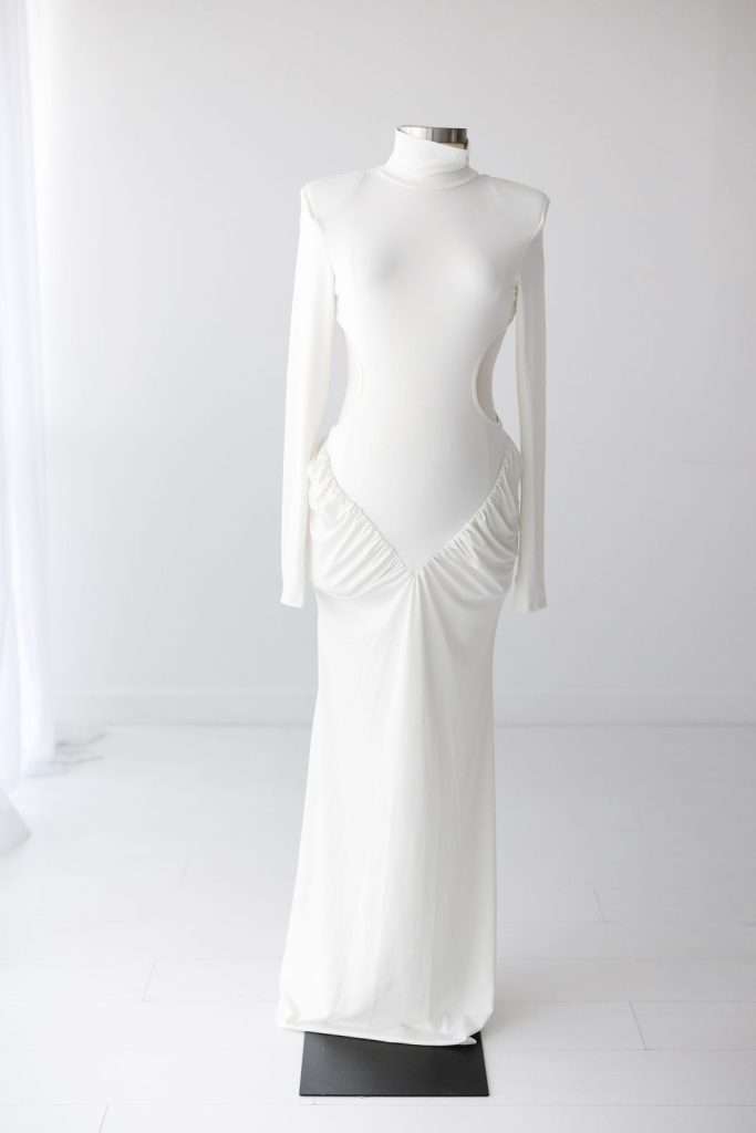 A white long-sleeve, floor-length gown with side cutouts and ruched detailing at the hips displayed on a dress form against a plain background.