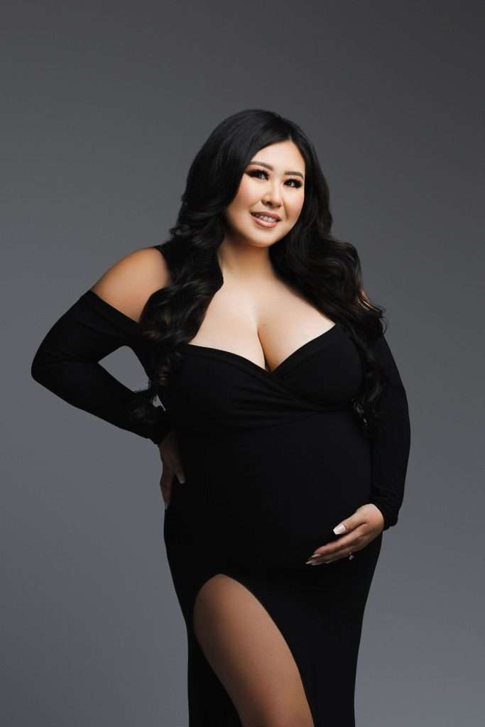 A pregnant woman in a black dress with long sleeves and a thigh-high slit poses against a plain gray background, smiling and holding her belly—a beautiful example of studio maternity photography Phoenix style.