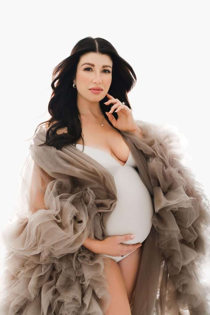 Pregnant woman in a sheer, ruffled robe holding her belly and looking at the camera against a white background, beautifully captured by a Family Photographer Phoenix.