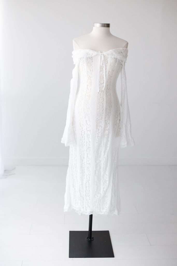 A white off-the-shoulder lace dress is displayed on a dress form against a plain white background.