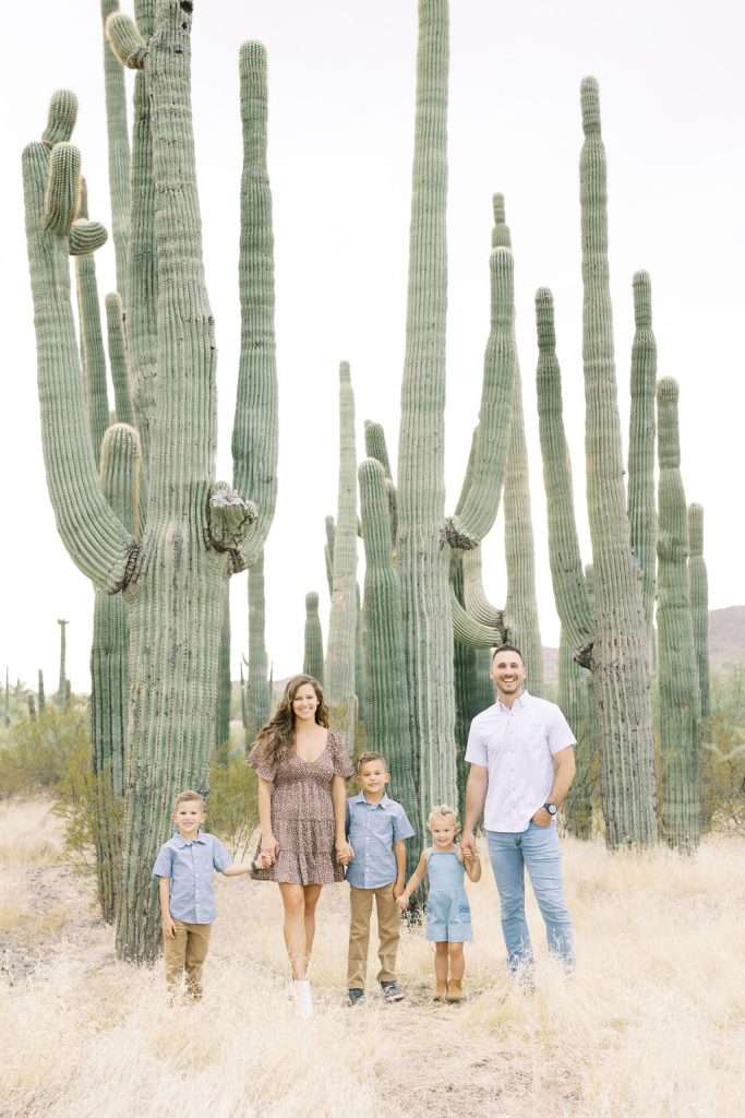 A family of five stands in a desert landscape among tall cacti, all dressed in light blue and neutral-toned clothing, captured beautifully by a Family Photographer Phoenix.