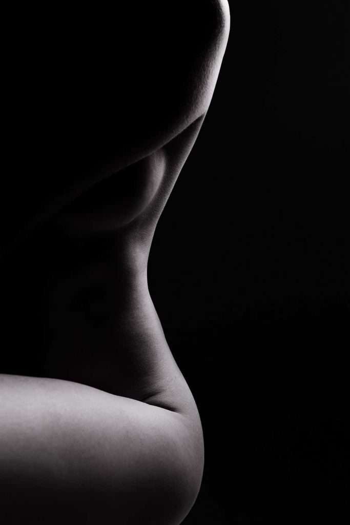 Black and white photo of a nude human torso, showing the curves of the waist, hip, and thigh against a dark background with dramatic lighting.