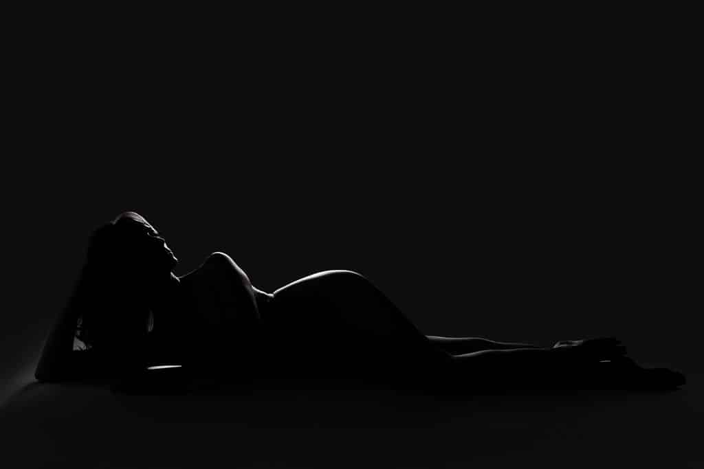 A pregnant woman is lying on her side in a dark setting, with her silhouette highlighted by soft backlighting.