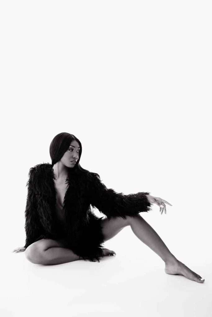 A woman in a black fur coat sits on the floor with one leg extended and one arm reaching out, against a plain white background.