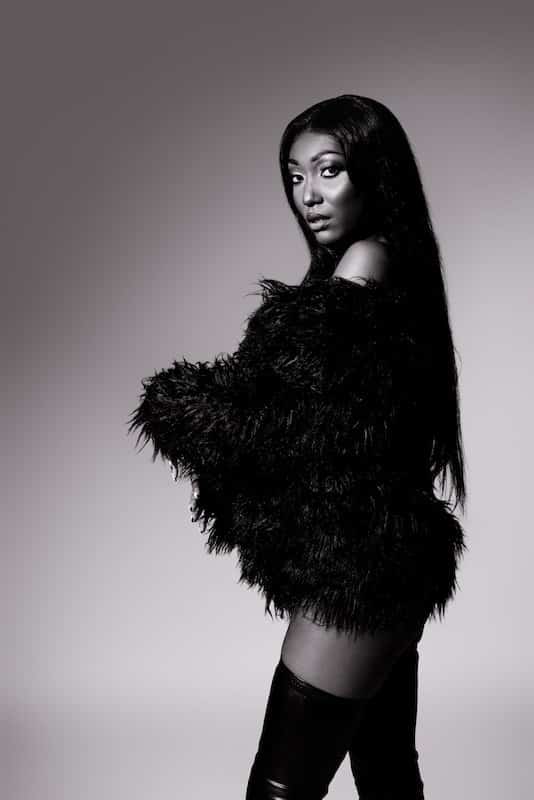 A woman with long hair poses in a fuzzy, off-the-shoulder black outfit and thigh-high boots against a plain background in a black-and-white photo.