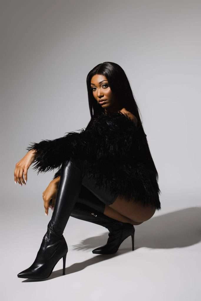 Woman with long straight hair squatting, wearing a black faux fur jacket and black knee-high heeled boots, posing against a plain light gray background.