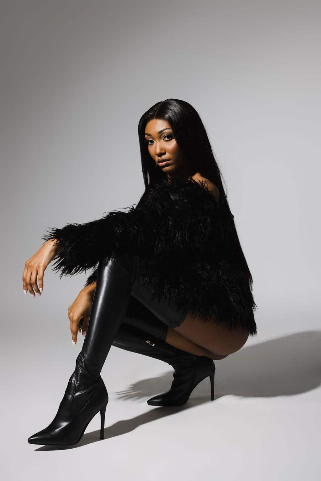 Woman with long straight hair squatting, wearing a black faux fur jacket and black knee-high heeled boots, posing against a plain light gray background.