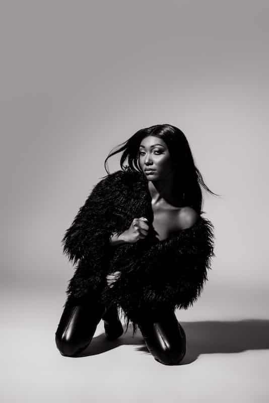 A woman wearing a black faux fur coat and shiny pants kneels on the floor in a studio with a plain background. The image is in black and white.