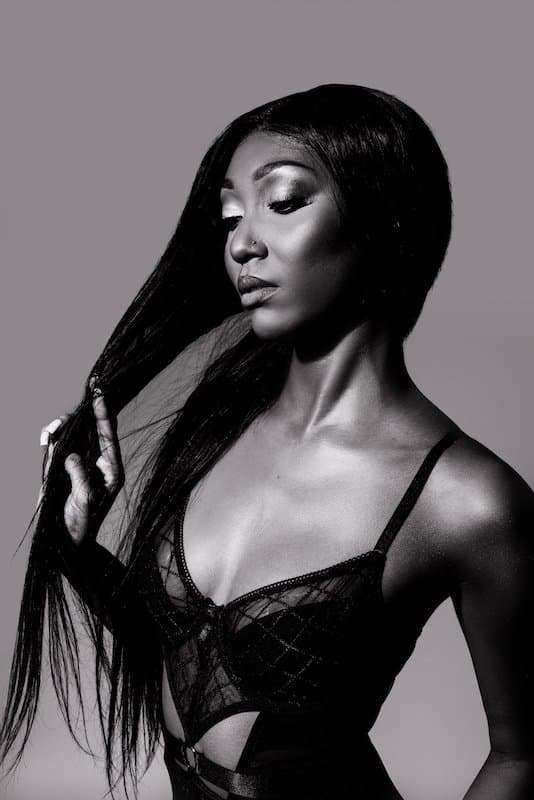 A woman in a black lace top poses against a plain background, holding a section of her long hair and looking down with a neutral expression.