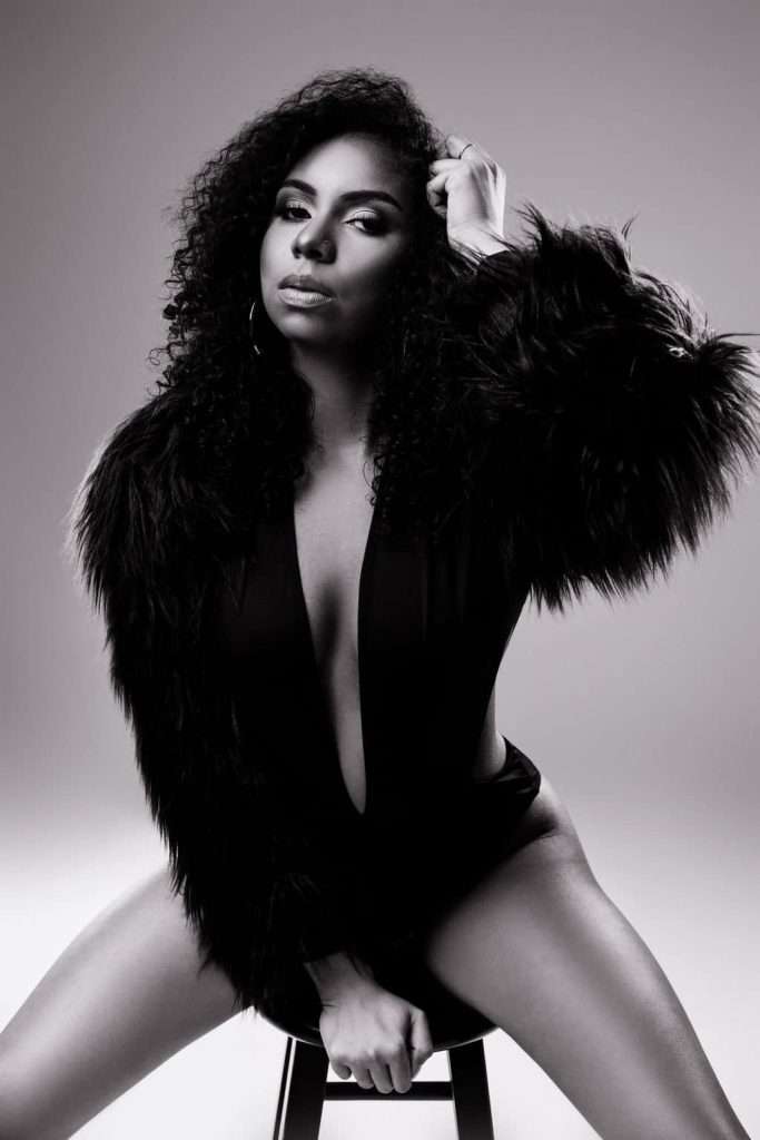 Black and white photo of a woman with curly hair wearing a fur jacket and bodysuit, sitting on a stool, looking directly at the camera.