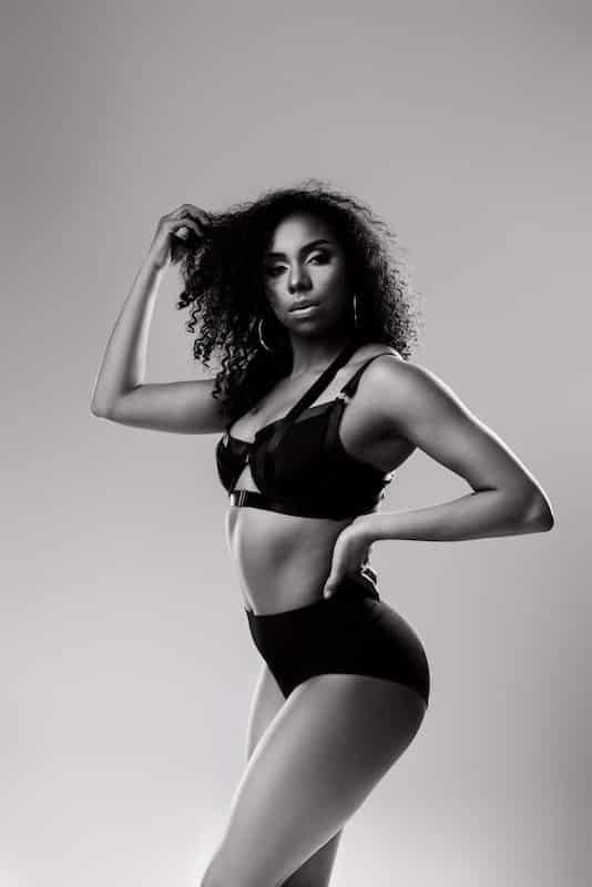 Black and white photo of a woman standing in profile, wearing a bra and high-waisted underwear, with one hand touching her curly hair.