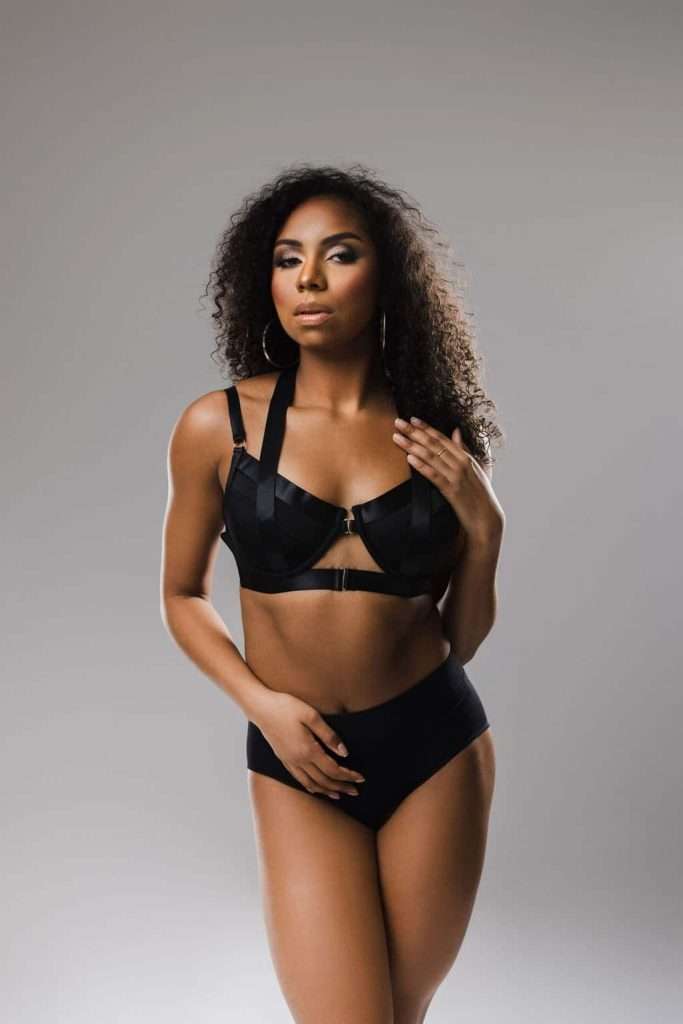 Woman with curly hair wearing a black bra and matching high-waisted underwear, standing against a plain gray background.
