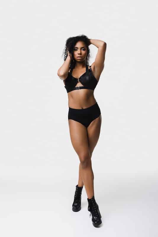 Woman standing in black lingerie and boots against a plain white background, with one hand on her head and the other on her hair.