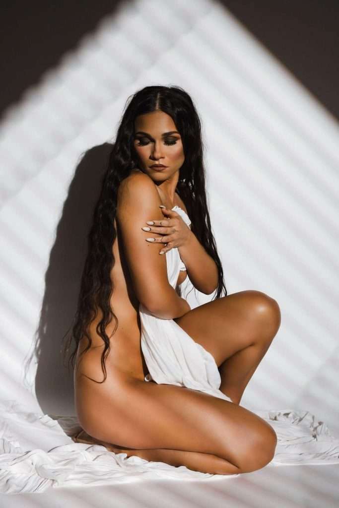 A woman with long dark hair sits on white fabric, partially draped in a white cloth, with diagonal shadows cast across her body and background.