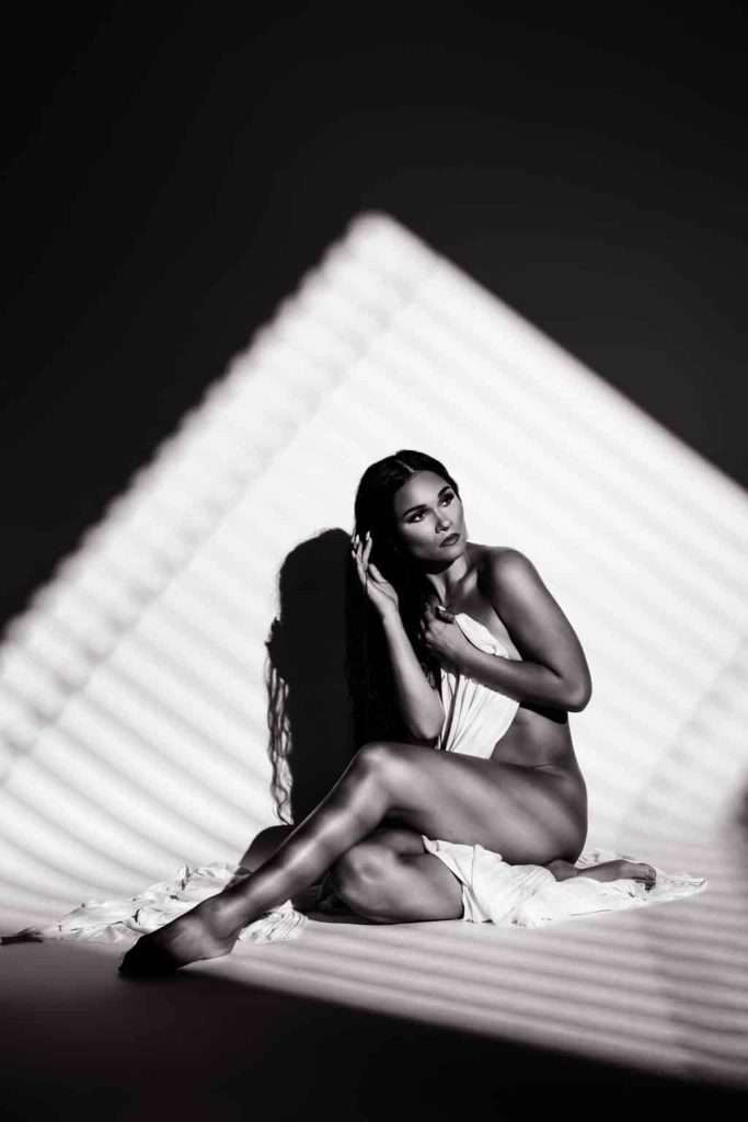 A woman sits on the floor draped in fabric, with striped light and shadow patterns cast across her body and background.
