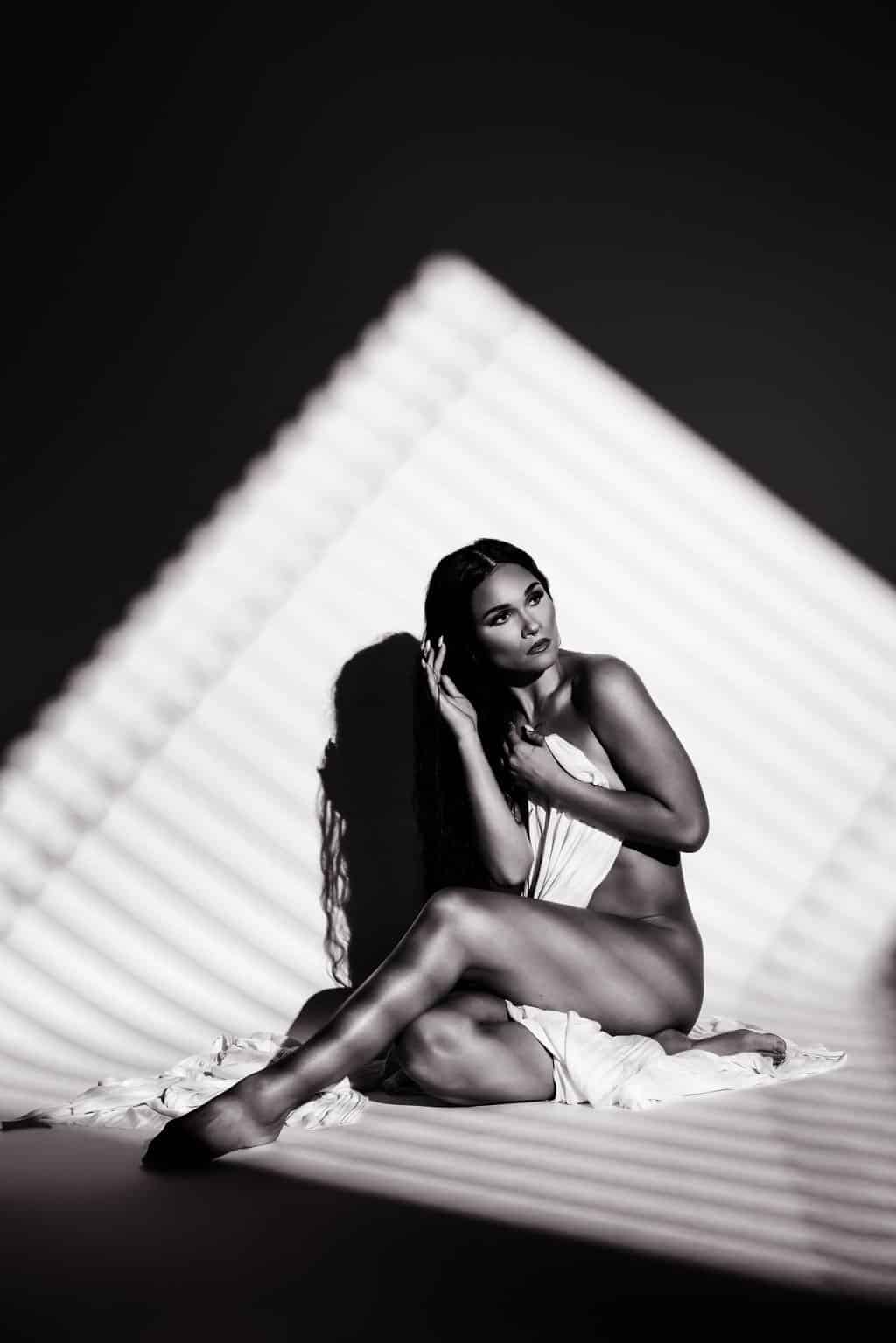 A woman sits on the floor draped in fabric, with striped light and shadow patterns cast across her body and background.