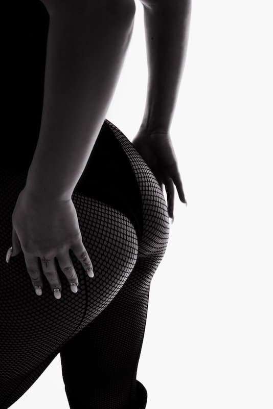 A person wearing fishnet tights and a bodysuit stands with one hand resting on their hip, shown from the waist down on a white background.