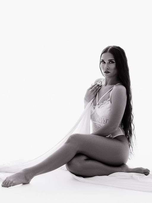 A woman with long hair poses seated on the floor, wearing a light lace outfit and holding a sheer fabric, against a plain light background.
