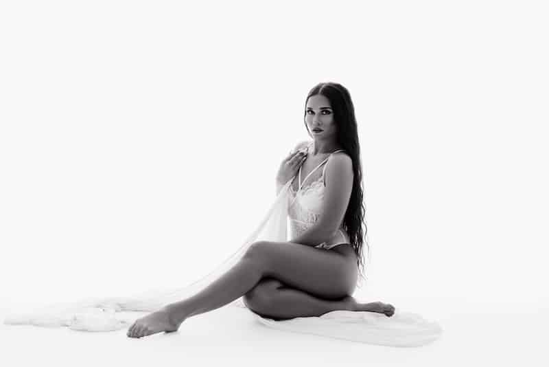 A woman with long hair sits on the floor in lingerie, draped with sheer fabric, against a white background.