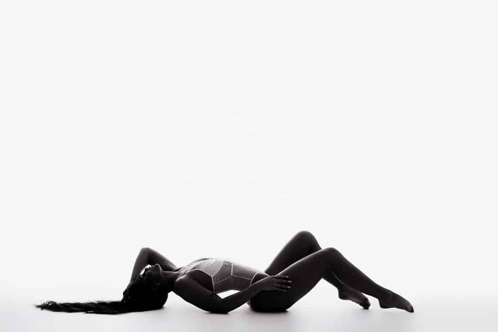 A woman in a bodysuit lies on her back on a white surface, with one knee bent and her long hair extended behind her. The image is high-contrast and minimalistic.