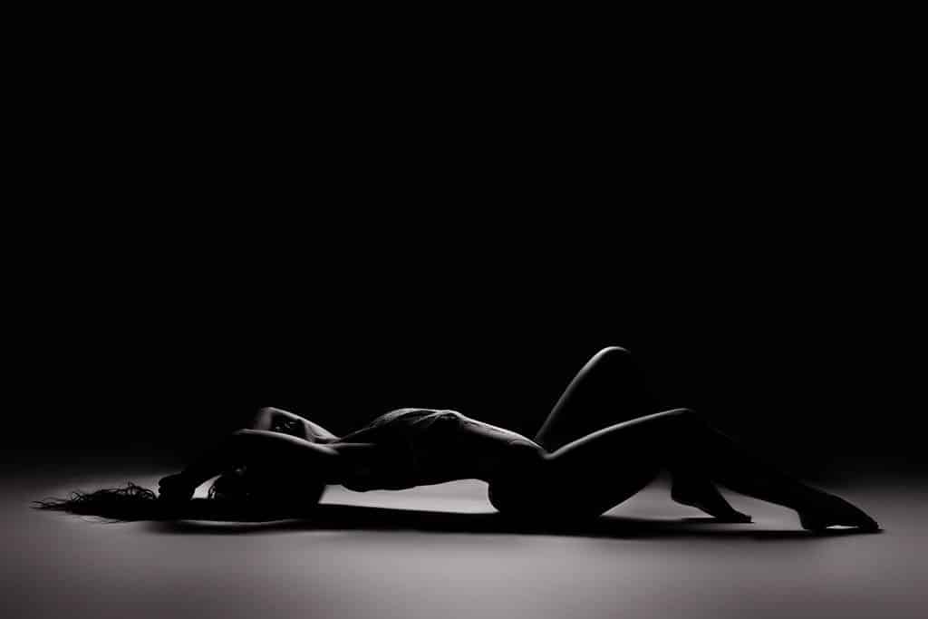 A person lies on their back with one leg bent and arms extended above their head, illuminated by dramatic side lighting against a dark background.