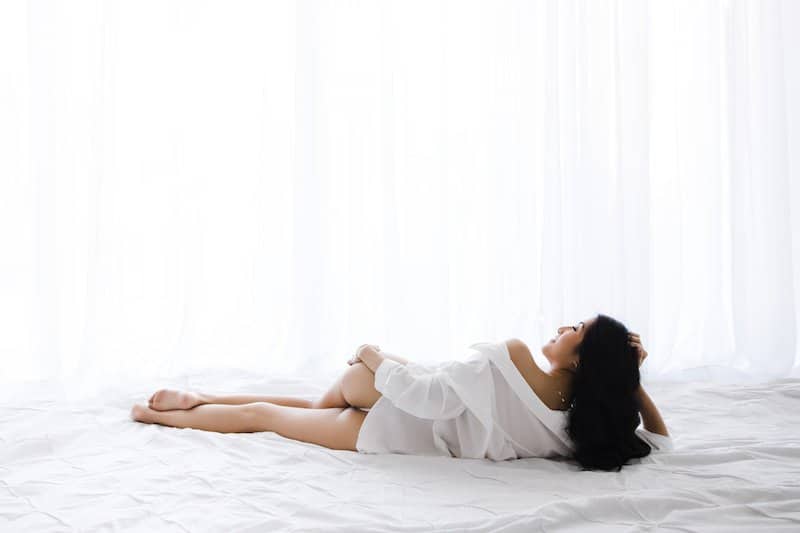 A woman in a white shirt lies on a white bed, facing to the side, with white curtains in the background.