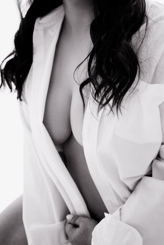 A person wearing an unbuttoned white shirt, partially exposing their chest, with long, dark hair visible; the image is in black and white.