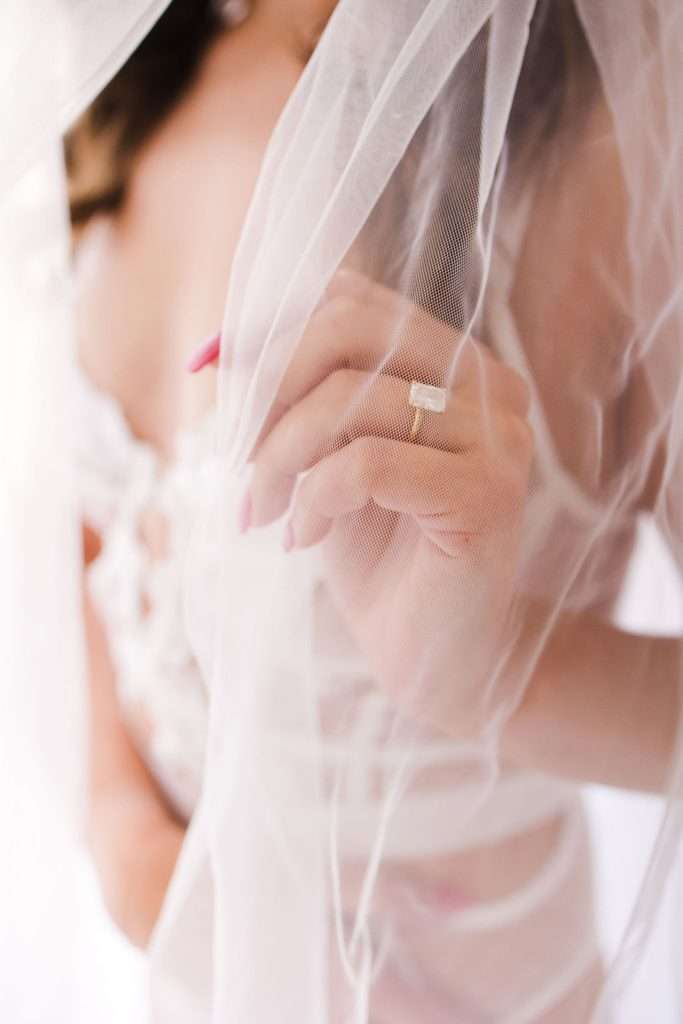 A person wearing a sheer bridal veil holds the veil with one hand, displaying a gold ring and manicured nails.