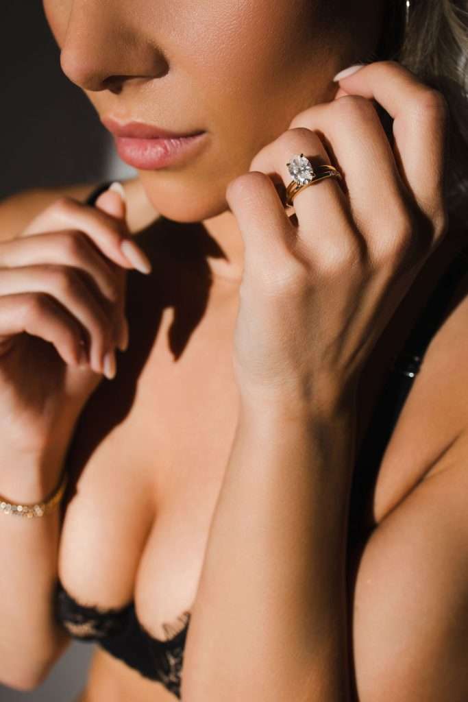 A woman wearing a black lace bra displays a large diamond ring on her finger, with her lips and hands in focus under natural light.
