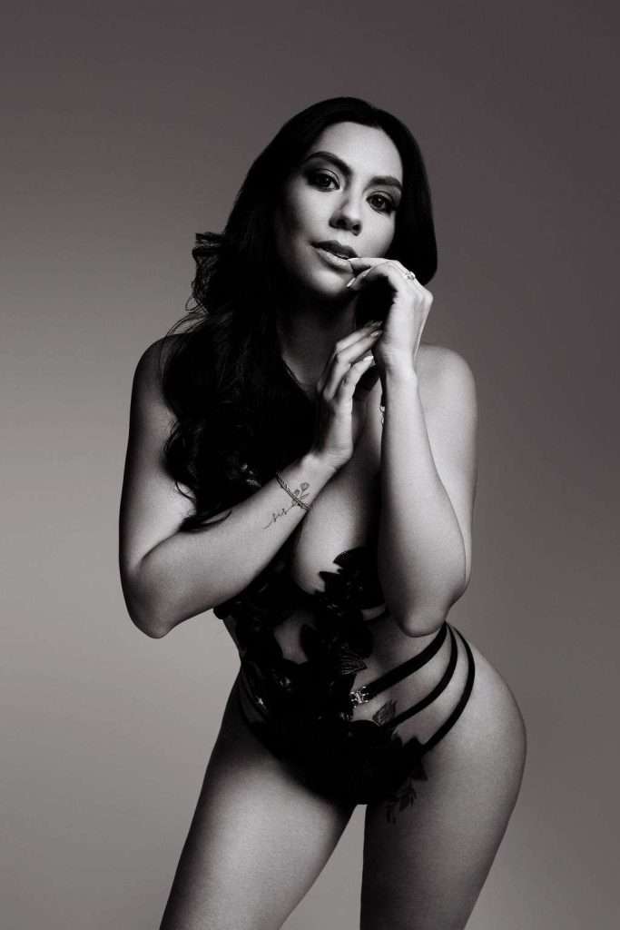 A woman with long dark hair poses in a revealing, strappy, black outfit against a plain, gradient background.