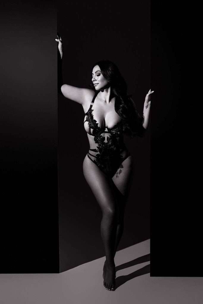 A woman poses barefoot in a black lace bodysuit against a dark background, with one arm raised and the other resting on a panel. The photo is in black and white.